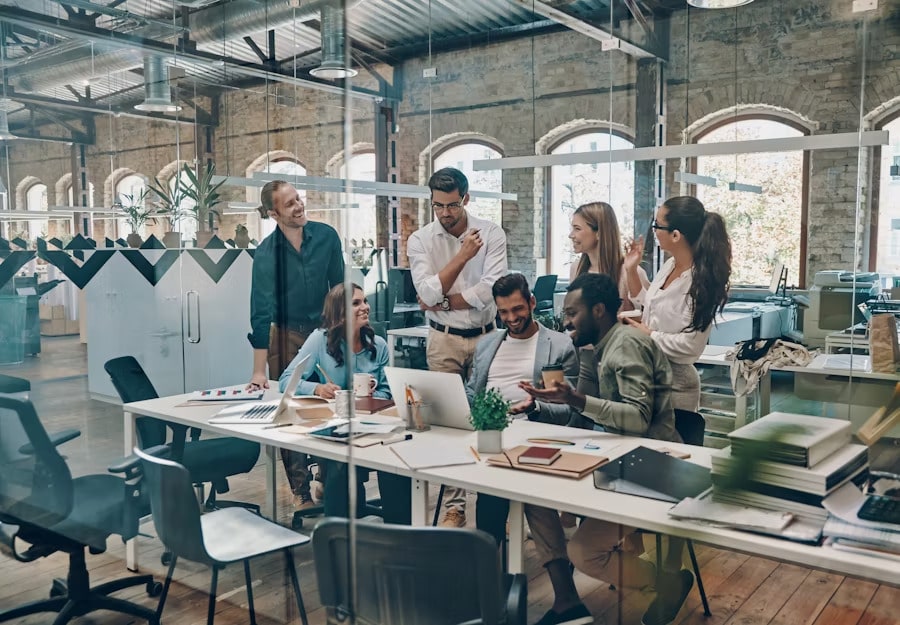 Un grupo de seis profesionales diversos reunidos alrededor de un escritorio en un espacio industrial convertido. Parecen estar mirando la pantalla de un portátil y compartiendo un momento de interacción o risa. La oficina presenta paredes de ladrillo expuesto, ventanas arqueadas y un techo industrial. El espacio de trabajo es moderno, con escritorios blancos y suministros de oficina visibles.