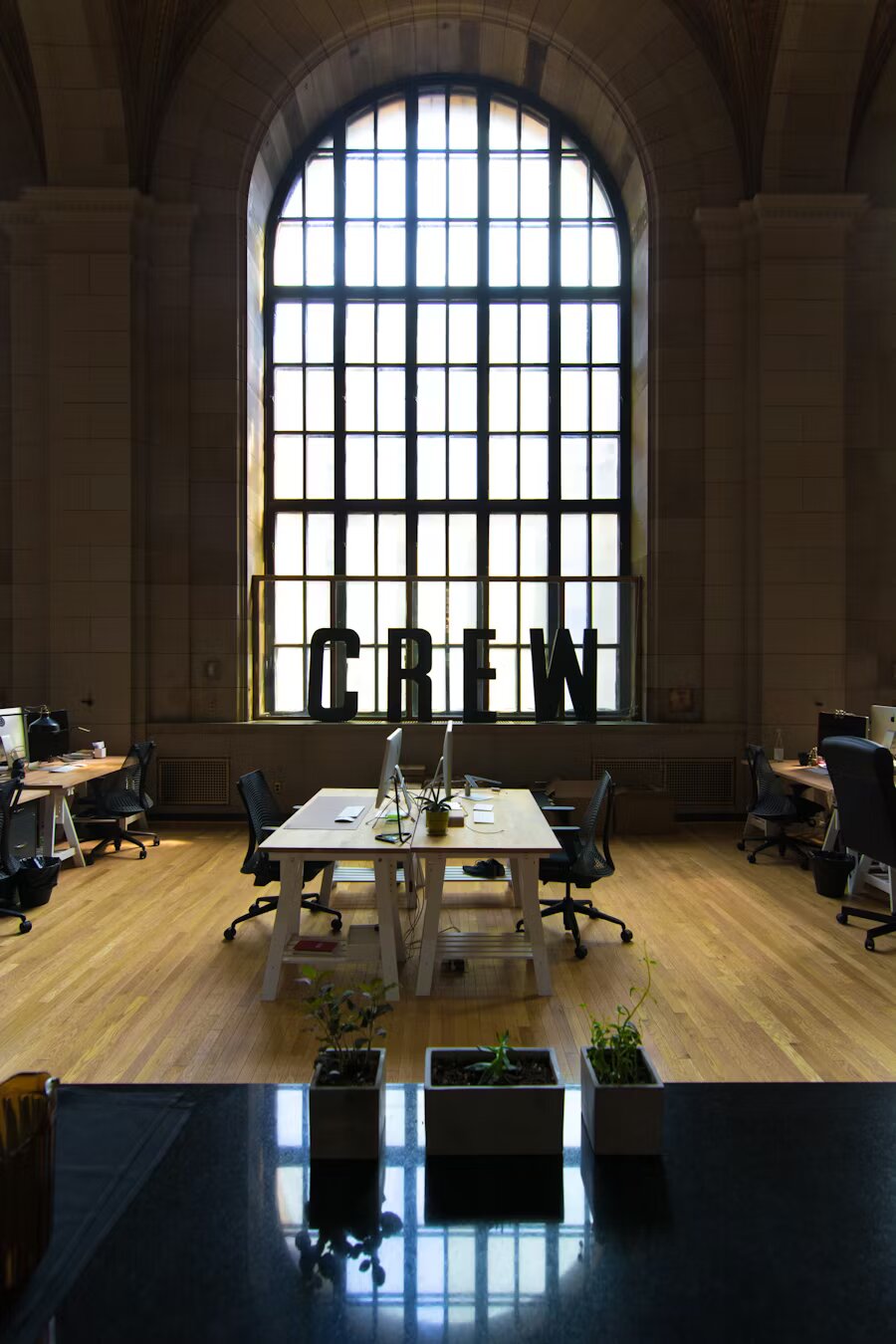 Espacio de coworking amplio y elegante con suelos de madera, escritorios compartidos y una gran ventana arqueada con la palabra “CREW” decorando el ambiente.