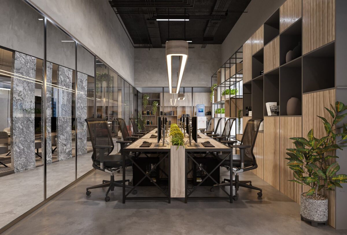 Estación de trabajo compartida en oficina moderna con escritorios de madera, sillas ergonómicas negras, plantas decorativas y estanterías abiertas, todo bajo una iluminación colgante contemporánea.
